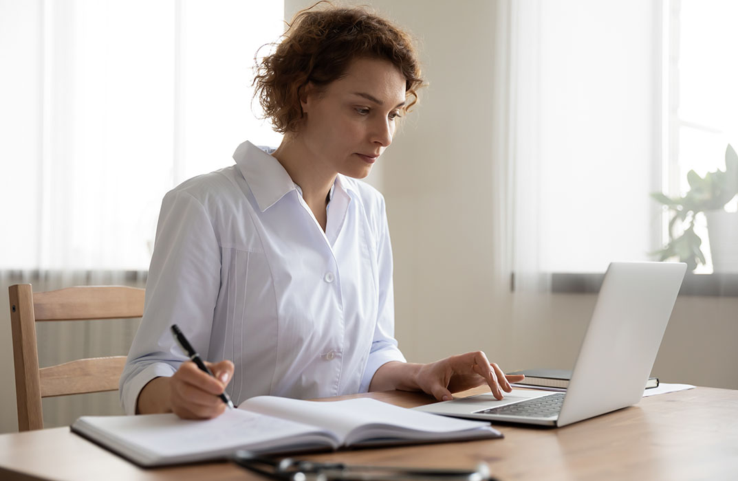 Young woman doctor working on laptop taking notes at workplace. Female physician writing in notebook using computer sitting at desk. Professional medic therapist everyday routine in hospital office.